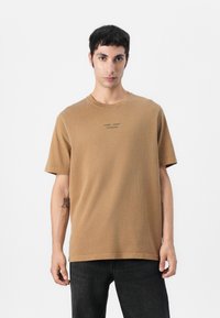 Camiseta de algodón marrón con un corte holgado, cuello redondo y mangas cortas. Presenta el texto impreso "SAMSØE SAMSØE COPENHAGUE" en el frente.