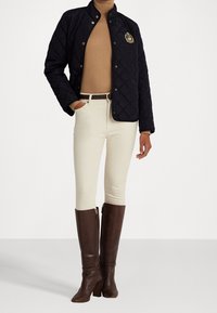 Tmavě modrá prošívaná bunda s vysokým límcem a zlatým emblémem, kombinovaná s béžovými přiléhavými kalhotami a hnědými kozačkami na podpatku až ke kolenům se zipy.