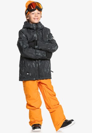 Schwarze Camouflage-Jacke mit Kapuze, kombiniert mit leuchtend orangen Schneehosen. Schwarze und orange Mütze, Skibrille mit verspiegeltem Glas, schwarze Stiefel.