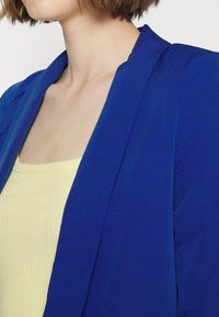Sininen bleiseri, jossa on sileä ja rakenteellinen muotoilu, notched-kaulus, yhdistettynä vaaleankeltaiseen ribattuun toppiin.