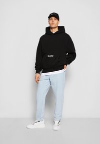 Czarna bluza z kapturem z przednią kieszenią i białym logo, w połączeniu z jasnoniebieskimi dresami i białymi sneakersami. Model stoi na tle jednolitym.