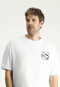 Camiseta de algodón blanca con mangas cortas, que presenta un gráfico de un patinete y el texto "Rider Conmigo" en el área del pecho izquierdo.