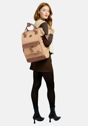 Junge Frau im schwarzen Kleid und Stiefeln, die einen großen hellbraunen und braunen Rucksack über eine Schulter trägt und über ihre Schulter zurückblickt.