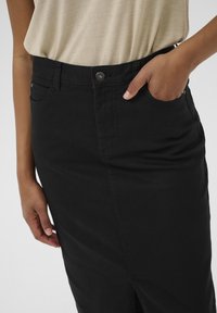 Jupe en denim noir au design ajusté, avec une fente à l'avant, une fermeture par bouton supérieur et des poches latérales, présentant une texture lisse et un léger stretch.
