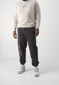 Felpa grigio chiaro con logo sul petto, abbinata a pantaloni della tuta grigio scuro con scritta bianca "OFF LIMITS". Scarpe da ginnastica bianche visibili.