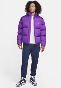 Giovane con capelli ricci che indossa un giubbotto imbottito Nike viola, una maglietta bianca, pantaloni cargo blu scuro e sneakers Nike bianche e blu.