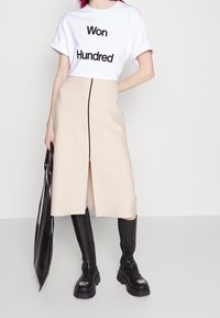 Camiseta blanca de manga corta con texto negro que dice "Won Hundred", combinada con una falda beige hasta la rodilla con una abertura frontal y cremallera negra. Las botas negras hasta la rodilla y un bolso de charol negro completan el atuendo.