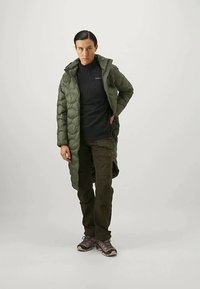 Zöld puffertakó, hosszú fazonú, kapucnival és steppelt textúrával, fekete hosszú ujjú felsőre és olíva zöld cargo nadrágra. Szürke cipőkkel viselve.