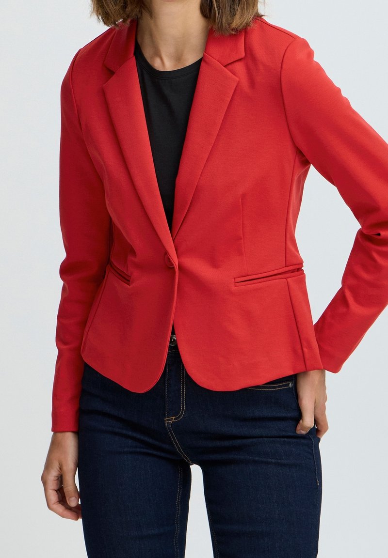 Blazer rosso con vestibilità sartoriale, realizzato in tessuto liscio, con colletto a revers, chiusura a un bottone e tasche laterali. Indossato sopra un top nero.