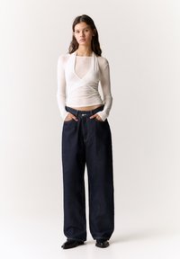 White, long-sleeve fitted top with a wrap design paired with oversized dark blue denim trousers featuring a front button closure and pockets.