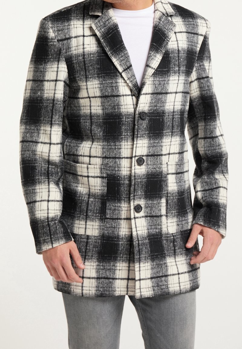 Homme portant un manteau à carreaux noir et blanc avec trois boutons sur une chemise blanche et un pantalon gris, les mains près des poches du manteau.