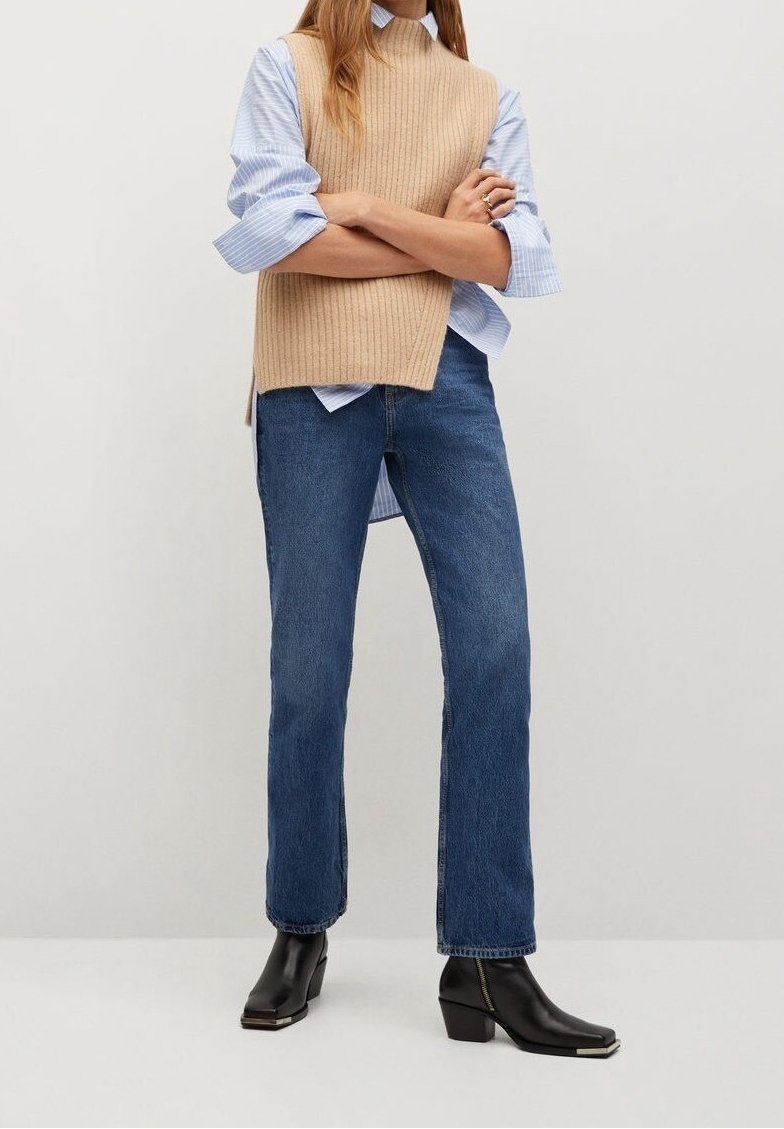 Femme portant un gilet en maille sans manches beige par-dessus une chemise rayée bleue, un jean bleu et des bottines noires, debout les bras croisés.