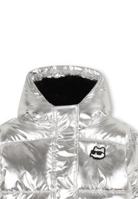 Sølv pufferjakke med hette, med en shiny finish, quiltet tekstur og svart pelskant. Inkluderer et katte-logo-lapp på brystet.