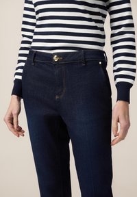 Jeans de mezclilla azul oscuro con pierna recta y detalles de botón dorado, combinados con un suéter a rayas en azul marino y blanco.