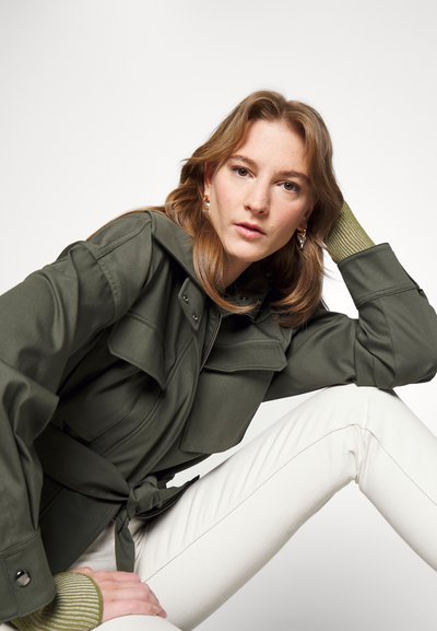 Casaco utilitário verde-oliva com cintura ajustada, grandes bolsos frontais e capuz. Combinado com calças justas brancas e punhos verdes canelados.