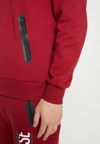 Czerwona bluza z ribbowanym dołem i mankietami, z czarną kieszenią na zamek po boku. Gładka faktura materiału.