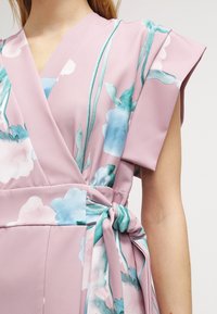 Robe portefeuille rose en tissu léger avec des motifs floraux bleus et verts, dotée de manches courtes et d'un détail de ceinture à nouer.