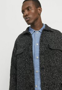 Veste en laine texturée noir et blanc avec une fermeture à bouton, dotée de deux poches poitrine, portée par-dessus une chemise en denim bleu clair.