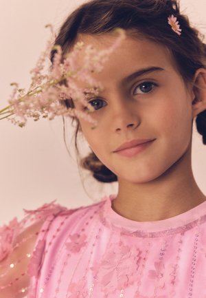 Młoda dziewczynka z ciemnymi zaplecionymi włosami i różowym kwiatowym spinką, ubrana w różową sukienkę z kwiatowymi aplikacjami, trzyma delikatne różowe kwiaty blisko twarzy.