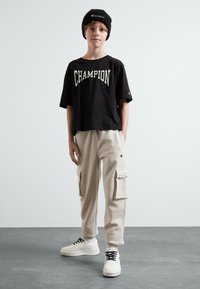Svart bomulls t-skjorte med "Champion"-logo, beige cargobukser med sidelommer, og hvite sneakers med svarte snøringer. Barnet har på seg en svart beanie.