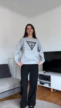 Grijze trui met een zwart driehoekig logo, gecombineerd met zwarte wijde broek. De stof lijkt zacht; ontspannen pasvorm met verlaagde schouders.