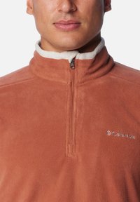 Fleece-Pullover in Rostfarbe mit einem Reißverschluss-Kragen, ausgestattet mit einem weichen cremefarbenen Innenfutter und einem kleinen Columbia-Logo auf der linken Brust.