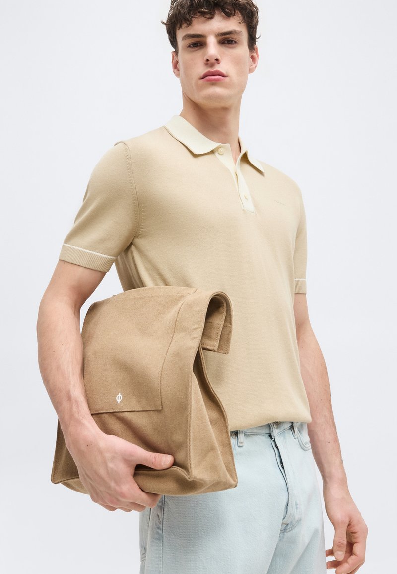Jeune homme portant un polo beige à manches courtes et un jean clair, tenant une veste en daim beige pliée, sur un fond clair uni.