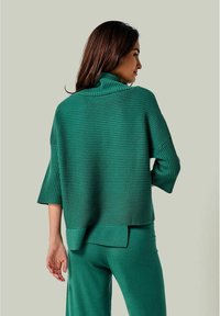 Grüner Strickpullover mit hohem Kragen, gerippter Textur, weiten Ärmeln und lockerer Passform. Kombiniert mit passender grüner Unterbekleidung.