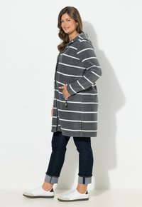 Cappotto a righe grigio con linee bianche, cappuccio, tasche frontali e cerniere. Indossato con jeans scuri e sneaker bianche. Tessuto dalla superficie liscia.