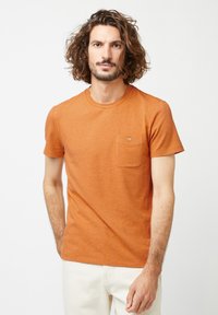 Korte mouwen, oranje T-shirt van textuurstof met een borstzak met een knoopdetail. Model draagt lichte broek.