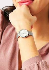 Orologio argento con quadrante rotondo, cinturino in acciaio inossidabile a maglia e un quadrante bianco con indicatori delle ore in cristallo. Indossato su un polso.
