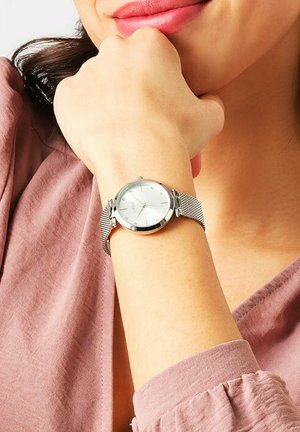 Silberne Uhr mit rundem Zifferblatt, mesh Edelstahlband und einem weißen Zifferblatt mit Kristall-Stundenmarkierungen. Am Handgelenk getragen.
