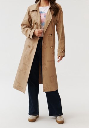 Vrouw draagt een beige trenchcoat met hondenschetsen en tekst, wit grafisch T-shirt, donkere wijde broek en lichte sneakers.