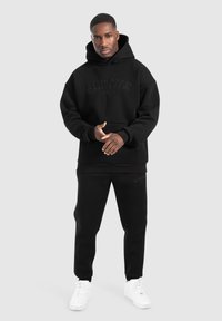 Černá mikina s kapucí a souprava tepláků vyrobená z měkké, texturované tkaniny. Na hrudi má vystouplé logo a odpovídající branding na kalhotách. Bílé tenisky.