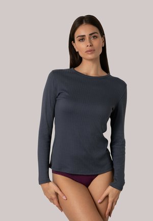 Donna con lunghi capelli scuri che indossa una maglia a maniche lunghe a coste di colore scuro e biancheria intima viola scuro, in piedi contro uno sfondo chiaro uniforme.