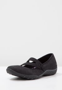 Zapatos planos negros con una parte superior texturizada, dos correas elásticas en el empeine y una suela de goma flexible.