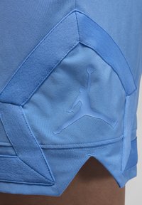 Blå atletiske shorts med glat stof, en tekstureret blå detalje ved kanten og en fremskudt logo-silhuet af en springende figur.