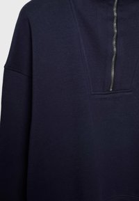 Sweat-shirt bleu marine à enfiler avec une fermeture éclair argentée partielle à l'avant et des détails visibles de couture autour de la fermeture éclair et de l’épaule.