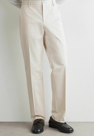 Pantalon beige sur mesure avec pli devant porté avec des chaussures en cuir noir à lacets sur un fond neutre de sol et mur.