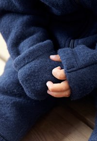 Enodelni termo pajac v mornarsko modri barvi z zadrgo, teksturirano tkanino, podvihanimi manšetami in tesnim prileganjem okoli rok.