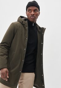 Olivgrüner, isolierter Parka mit abnehmbarem Kapuzen und mit Reißverschluss sowie Druckknöpfen, getragen über einem schwarzen, gestrickten Pullover.