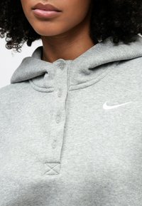 Γκρίζο φούτερ με κουμπιά στο μπροστινό μέρος, κουκούλα με κορδόνια, απαλό ύφασμα και λευκό λογότυπο Nike στο στήθος. Διαθέτει ραμμένη λεπτομέρεια σε σχήμα Χ.