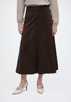 Jupe marron foncé mi-longue portée avec un haut beige à manches longues et des chaussures Mary Jane à talons beiges sur fond blanc.