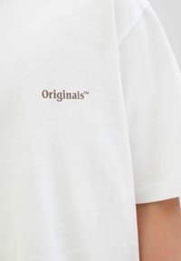 Valge puuvillane T-särk, millel on rinna peal väike pruun "Originals™" logo. Lihtne disain ümmarguse kaelusega ja lühikeste varrukatega.