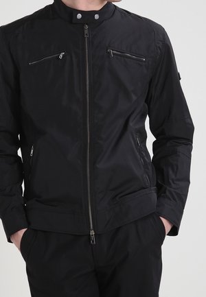 Mężczyzna ubrany w czarną kurtkę zapinaną na zamek błyskawiczny z kieszeniami na piersi i po bokach, stojący z rękami w kieszeniach spodni na jednolitym tle.