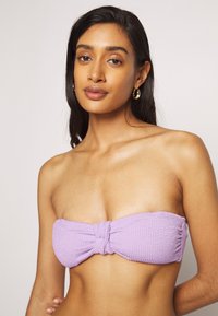 Top bandeau texturizado em tom de roxo com detalhe frontal em nó, feito de um tecido suave e sem alças, adequado para uso na praia.