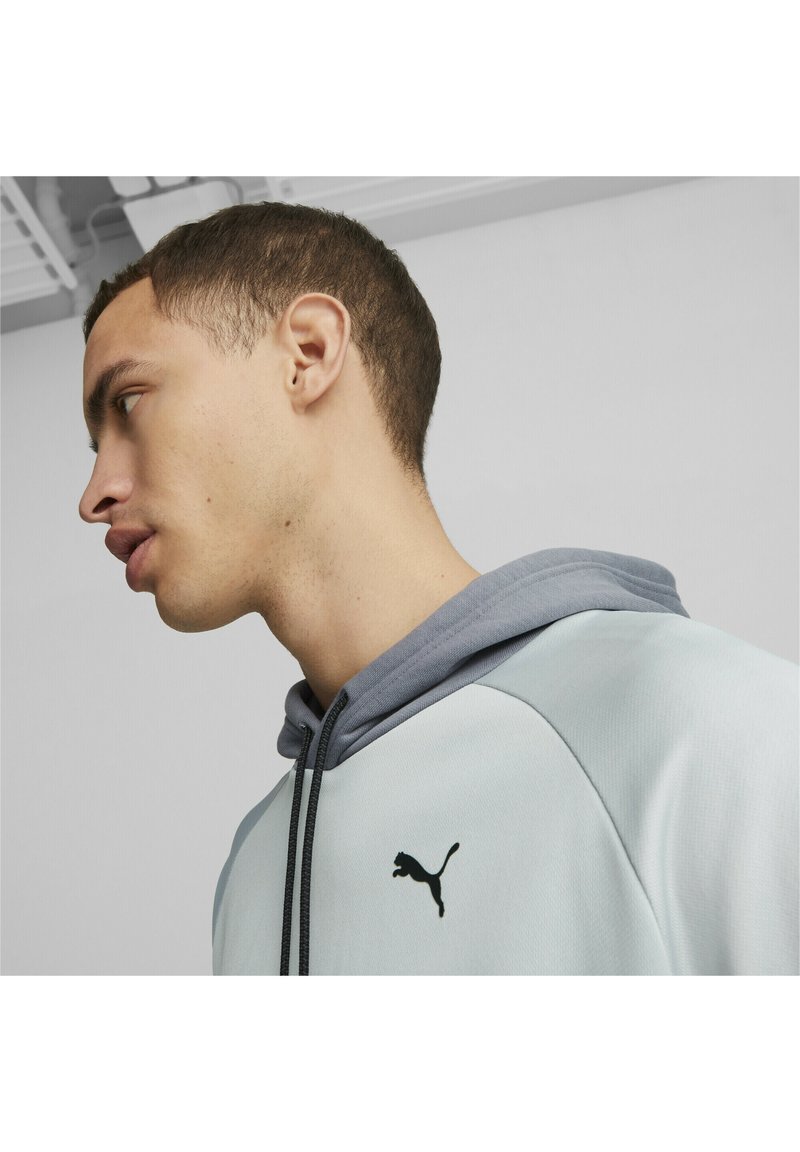 Felpa sportiva azzurro chiaro con cappuccio grigio, lacci neri e logo Puma nero sul petto. Tessuto liscio con un design aderente.