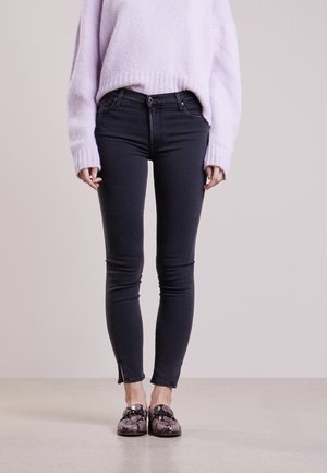Dark denim skinny jeans with a slim fit and ankle slits, paired with a light purple knit sweater and patterned slip-on shoes.