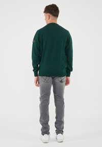 Maglione verde a maglia con colli rotondo, indossato sopra jeans grigi e sneakers bianche. Presenta una toppa sulla spalla sinistra.