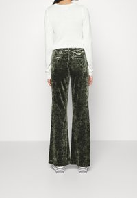 Grüne Samt-Hose mit weitem Bein und weicher Textur, kombiniert mit einem weißen Pullover. Sichtbare Gesäßtaschen und lässige Passform.